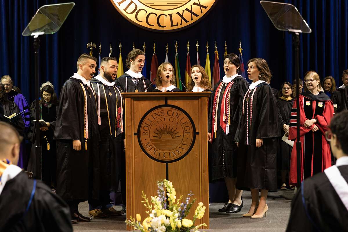 Students stand around a podium singing.