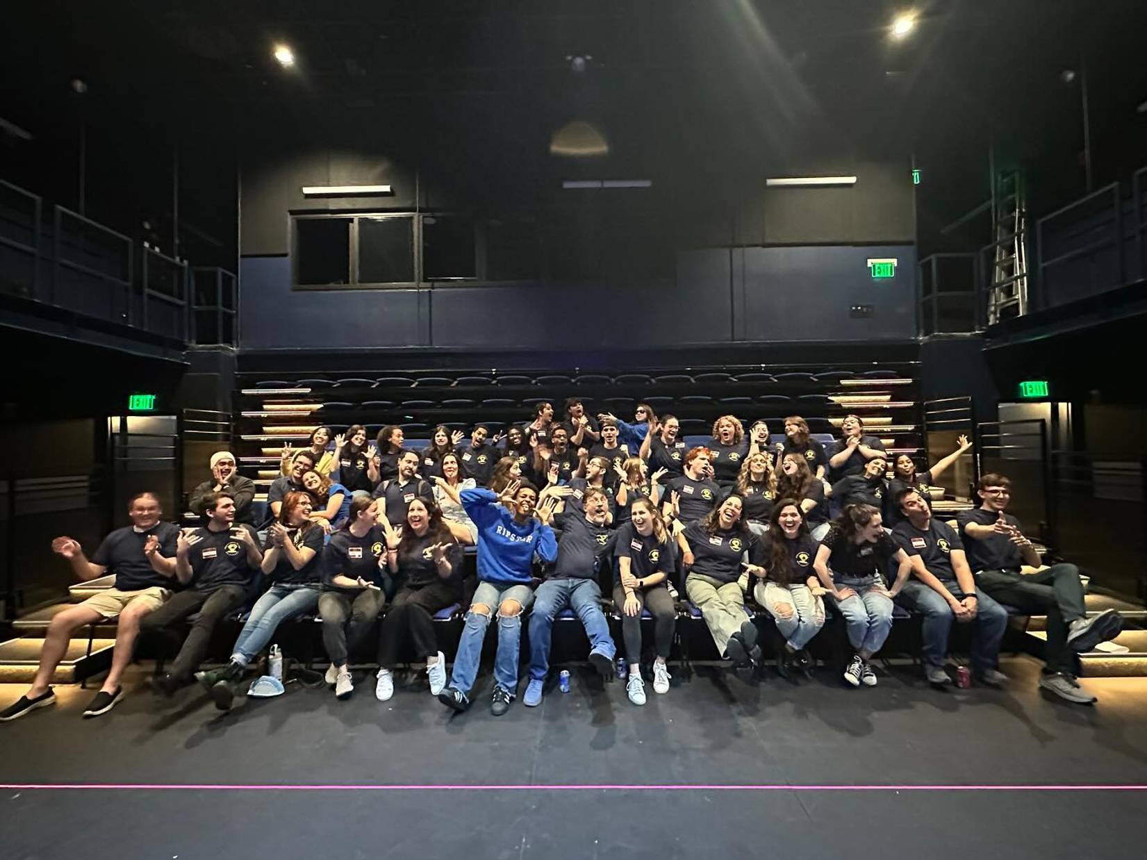 Large theater group posing playfully together onstage in matching shirts