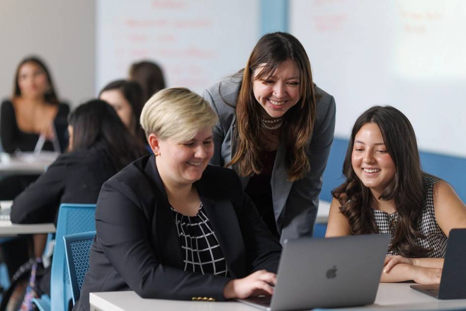 Smiling students and a professor collaborate around a laptop in a modern classroom setting.