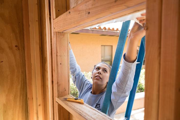 A student on a ladder reaching up to seal in the window of a tiny home class project.