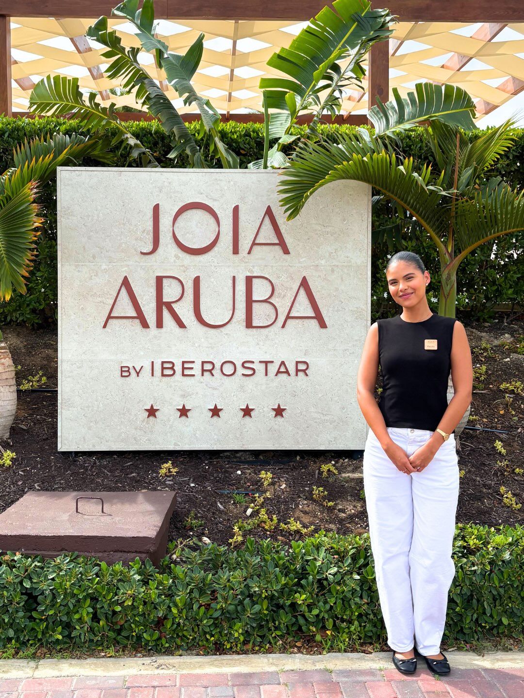 Student in a black top and white pants stands beside the "Joia Aruba by Iberostar" resort sign, framed by tropical plants.