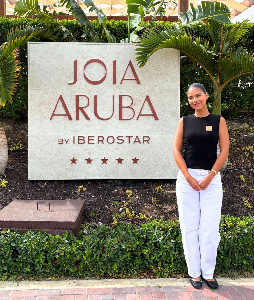 Student in a black top and white pants stands beside the "Joia Aruba by Iberostar" resort sign, framed by tropical plants.