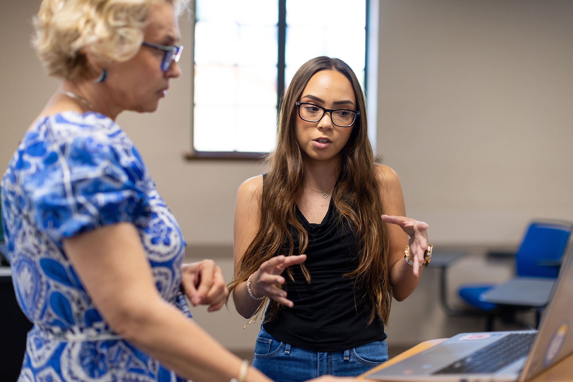 Student explains project on laptop to instructor in classroom setting.