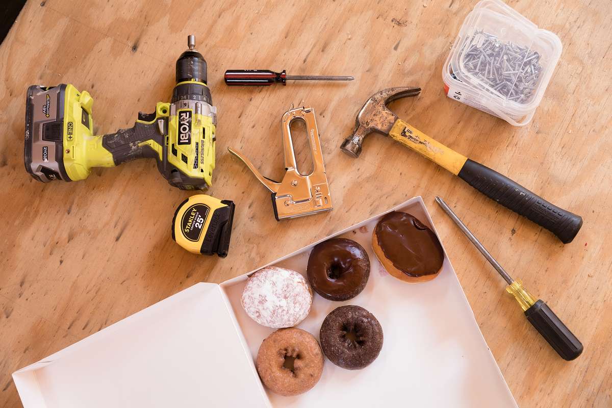 A selection of necessary tools laid out on the floor to build a tiny home: drill, screws, hammer, stapler, screwdrivers and of course a box of donuts.