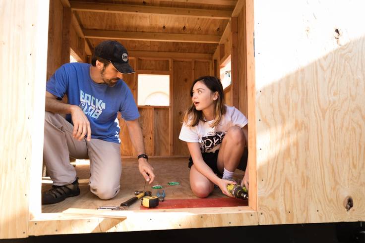 Professor Joshua Almond and college student kneeling in the doorway of a tiny home class project as she is using a drill to screw on a piece of plywood.