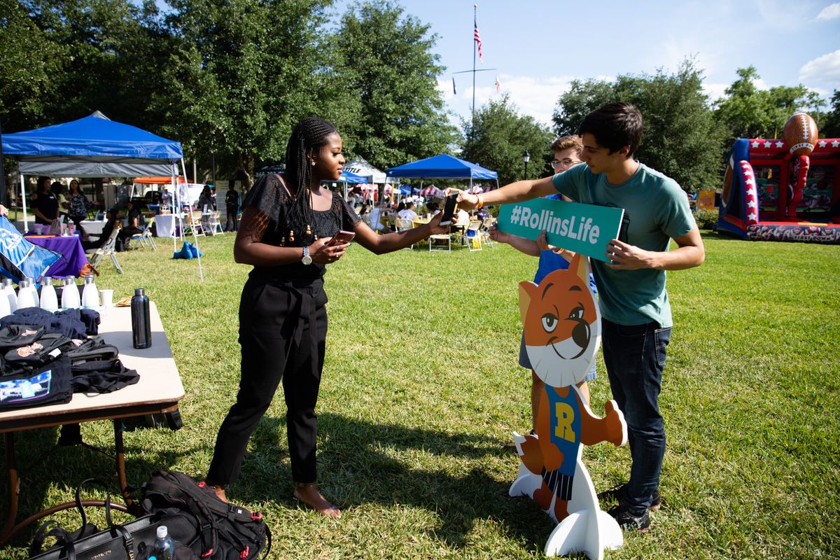 A British student on Rollins campus talking with two other students posing with a #RollinsLife sign and cutout fox.