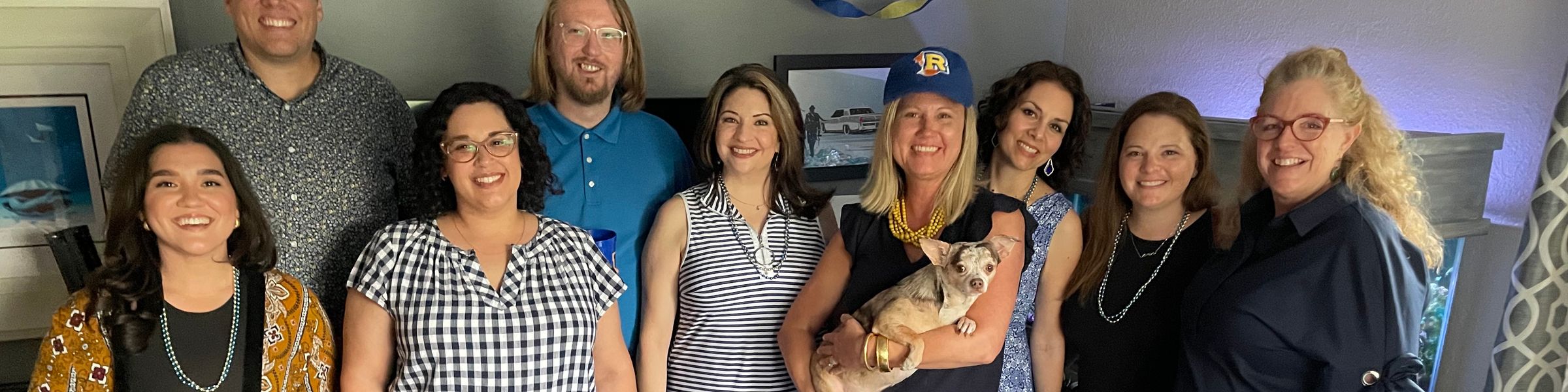 Group of alumni standing in a living room, one holding a dog.