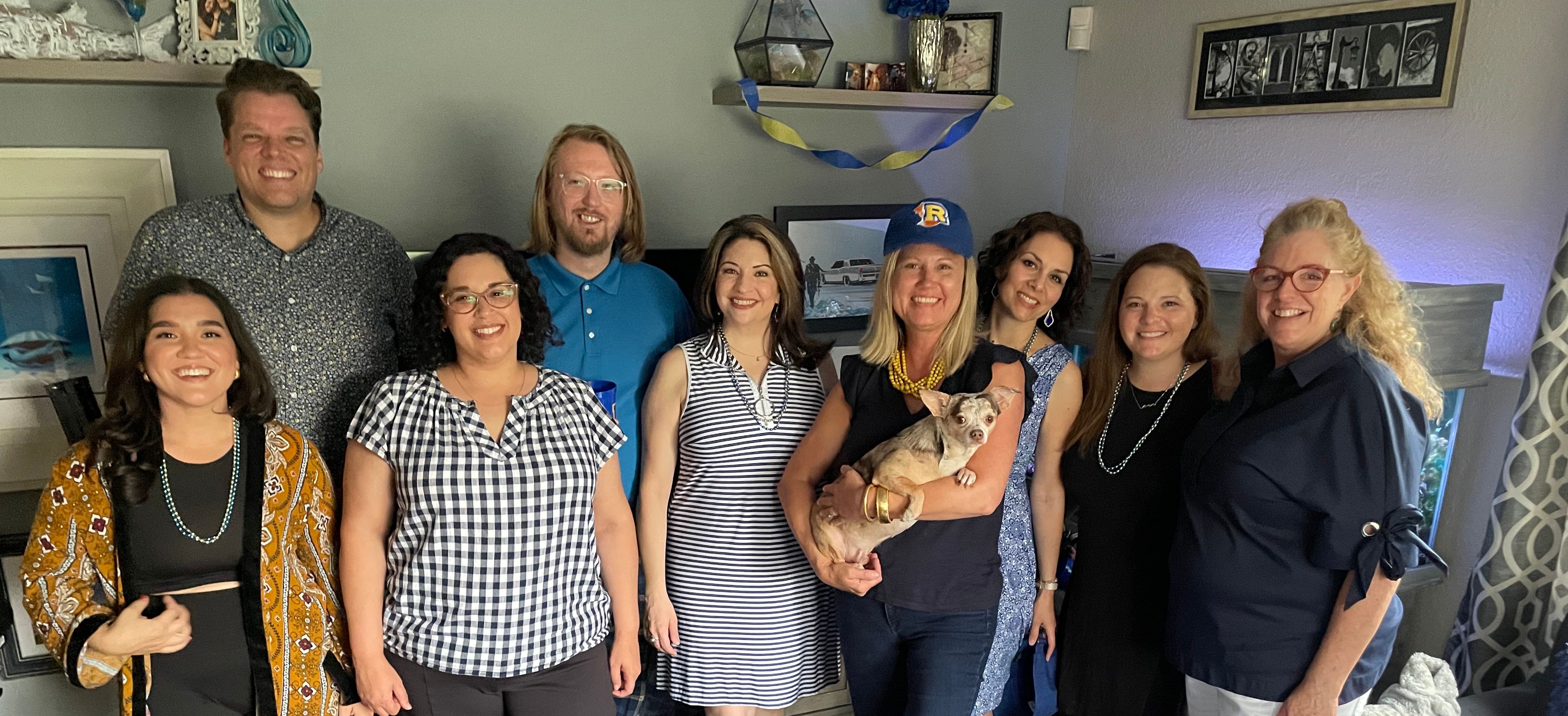 Group of alumni standing in a living room, one holding a dog.