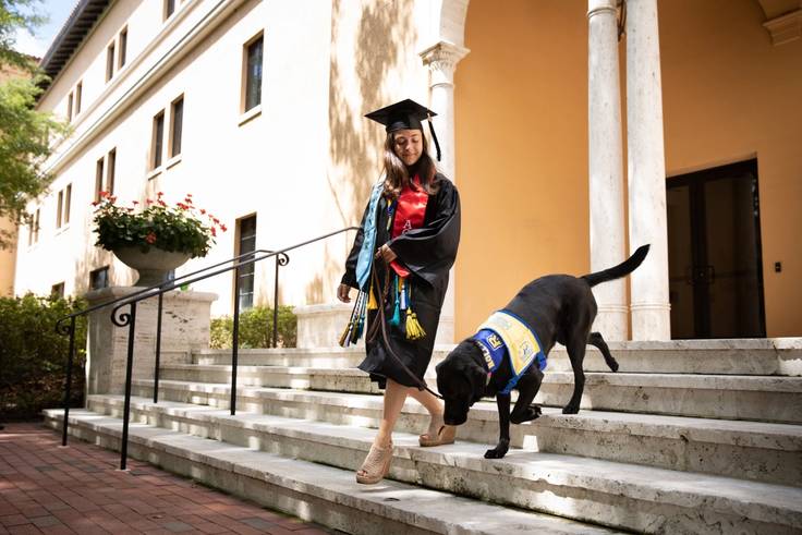 Marissa Cobuzio, in her commencement attire, walks down steps with a service dog.