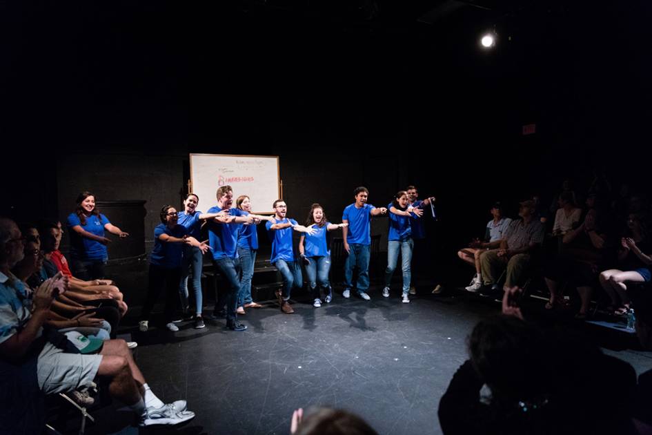 A line of students in matching blue t-shirts in front of an audience, arms outstretched