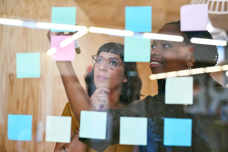 Two communications professionals brainstorm a content strategy using post-it notes on a glass board.