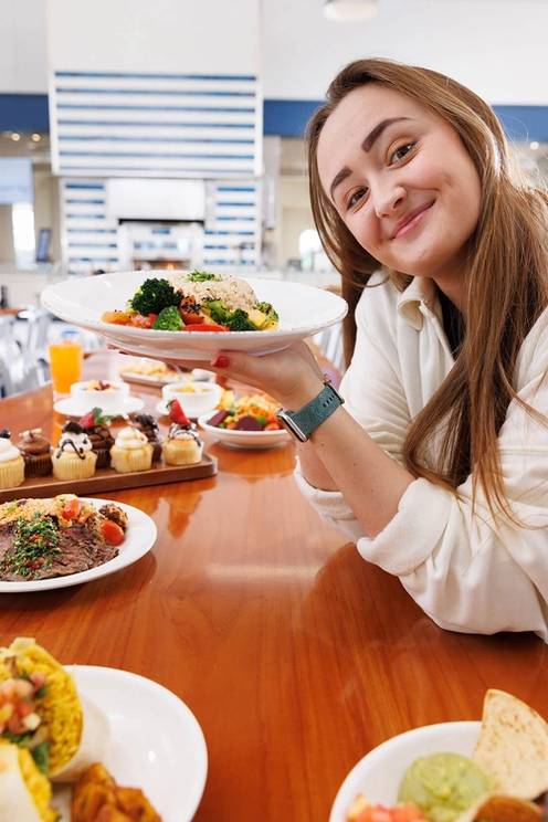 A student holds up a plate of food and is surrounded by other plates of food at the Marketplace.