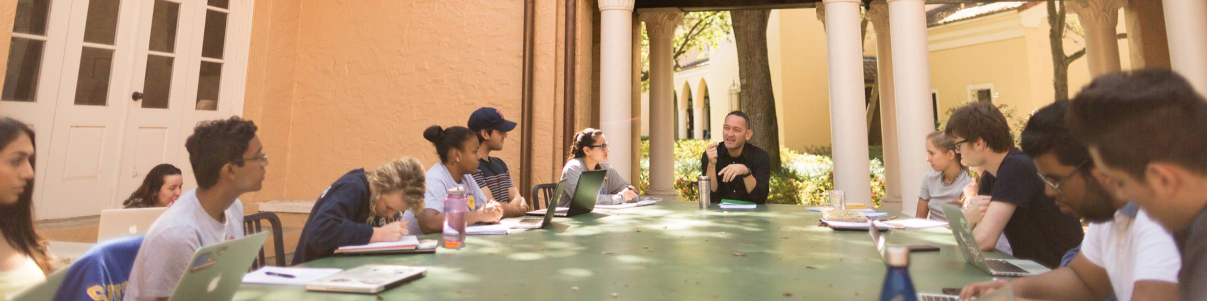 Rollins class being held in an outdoor classroom, a large table under oak trees, professor at the head and students on both sides