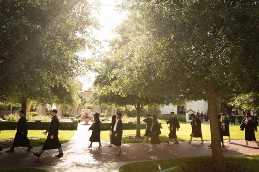 Students walk in cap and gown to a commencement ceremony.