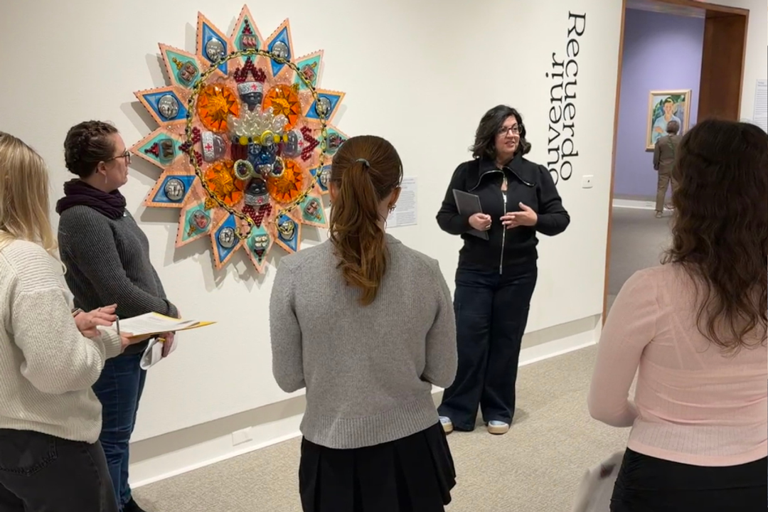 woman stands in front of an orange wall in an art gallery surrounded by people she is talking to