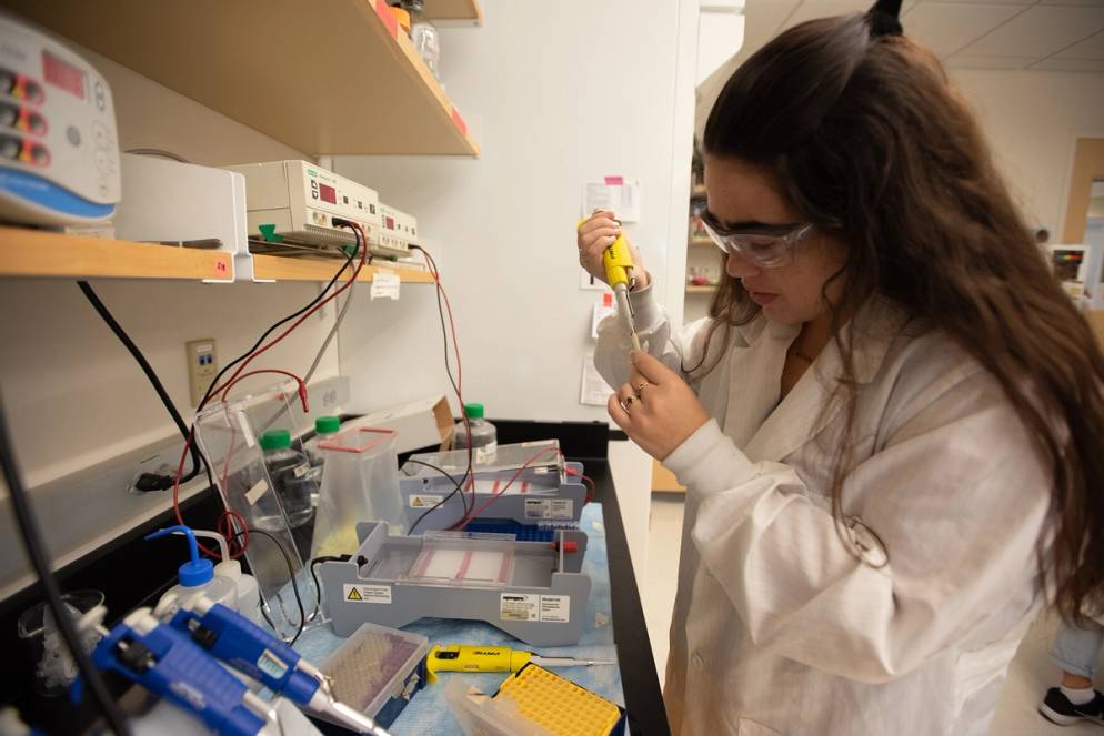 A student with a lab coat and protective glasses taking a DNA sample. 