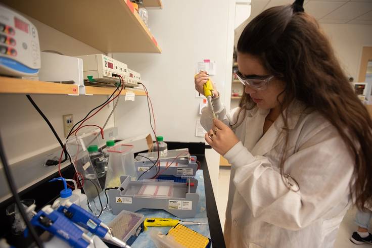 A student with a lab coat and protective glasses taking a DNA sample.