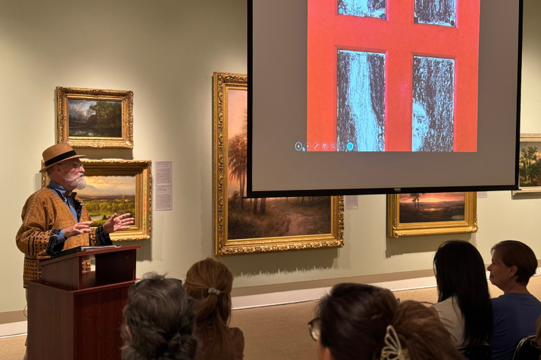 Bearded man stands at a lecturn talking. Red artwork is projected on a projection screen, crowd sits in an art gallery