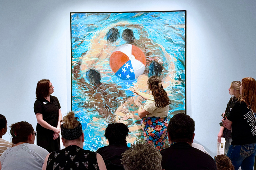 People sit and stand in an art gallery while a man points to a painting