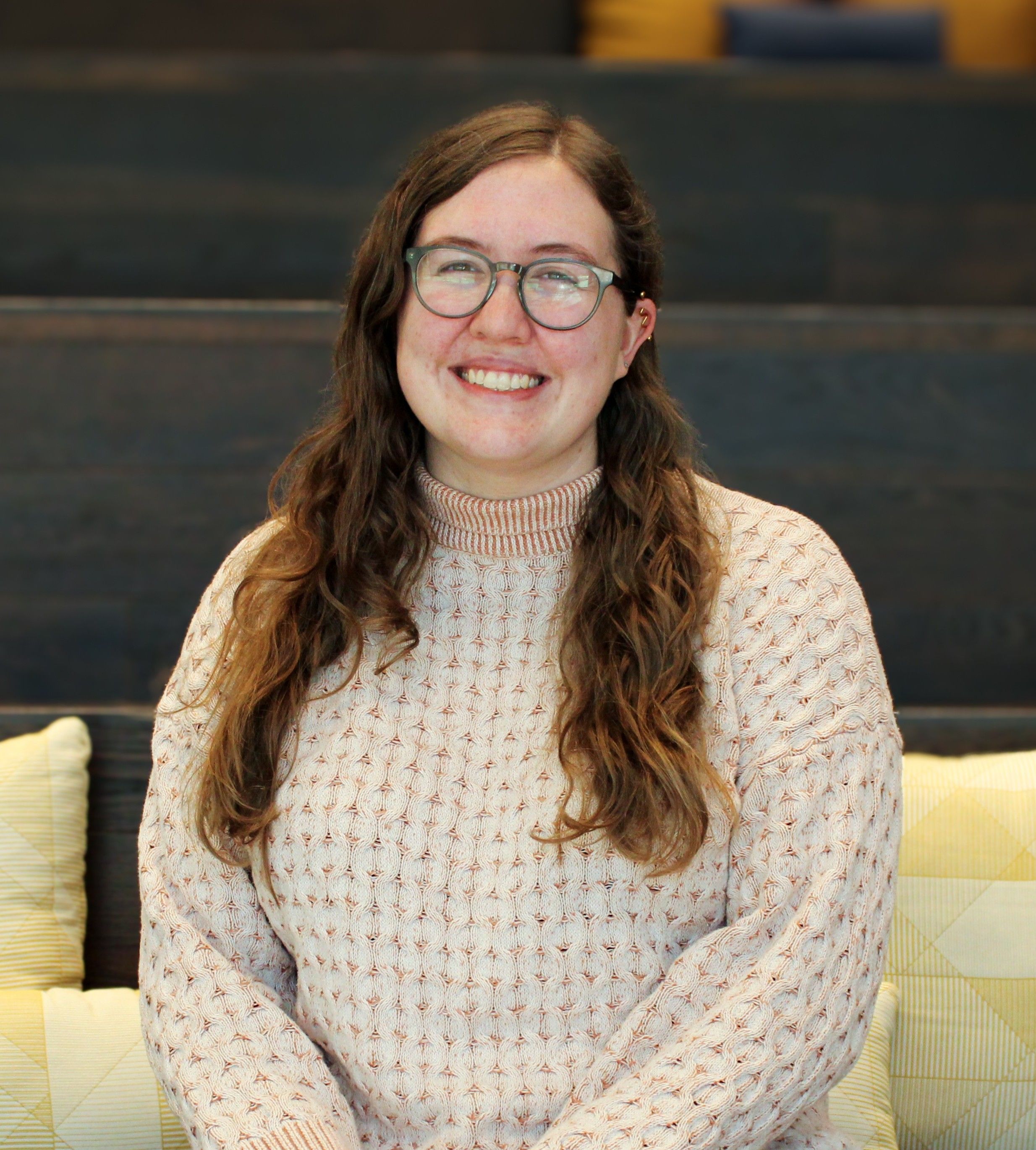 a picture of Cailey Cassity-Ness sitting on the staircase. She has brown curly hair, green glasses, and is wearing and orange and white sweater. 