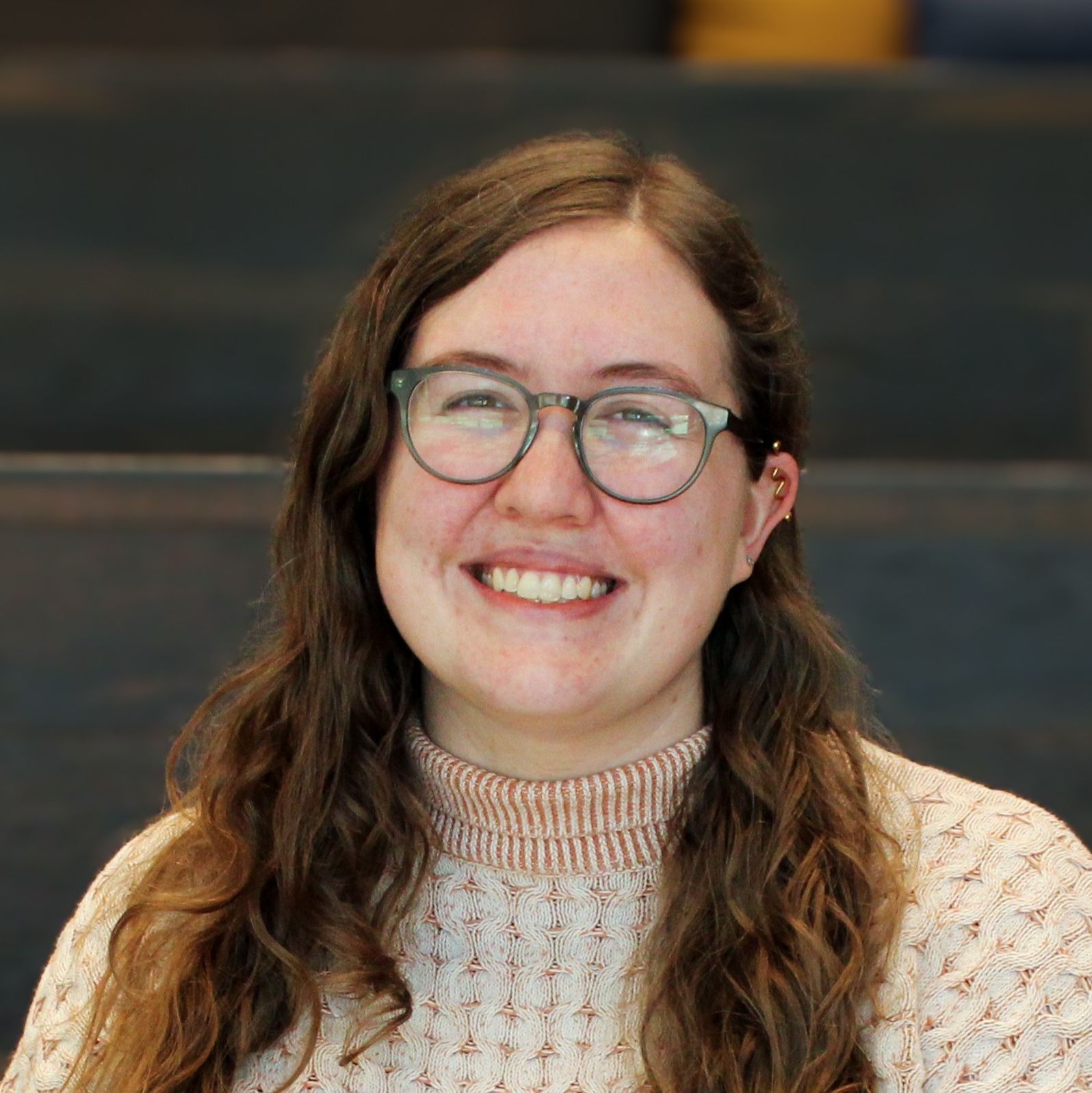 a picture of Cailey Cassity-Ness sitting on the staircase. She has brown curly hair, green glasses, and is wearing and orange and white sweater. 