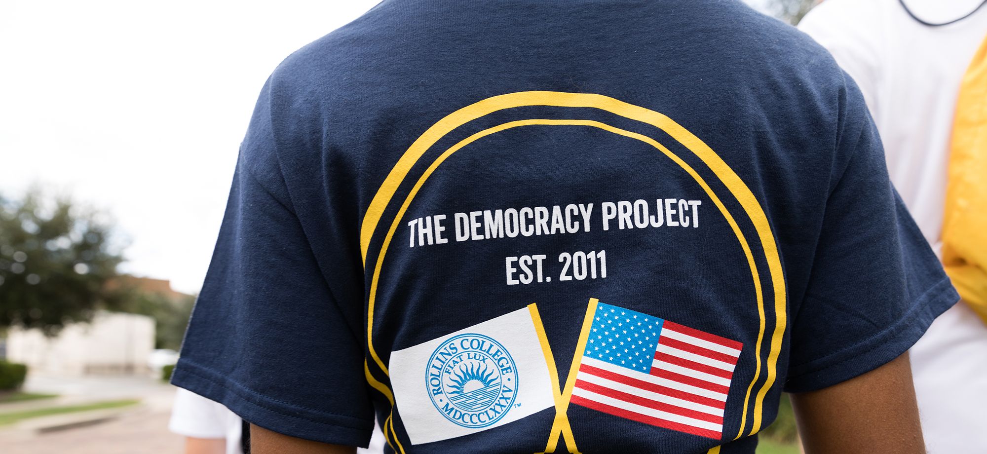 A student's back to see a t-shirt that says "The Democracy Project est. 2011"