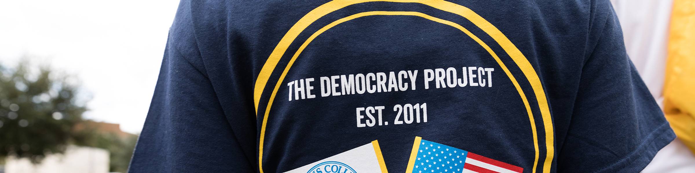 A student's back to see a t-shirt that says "The Democracy Project est. 2011"