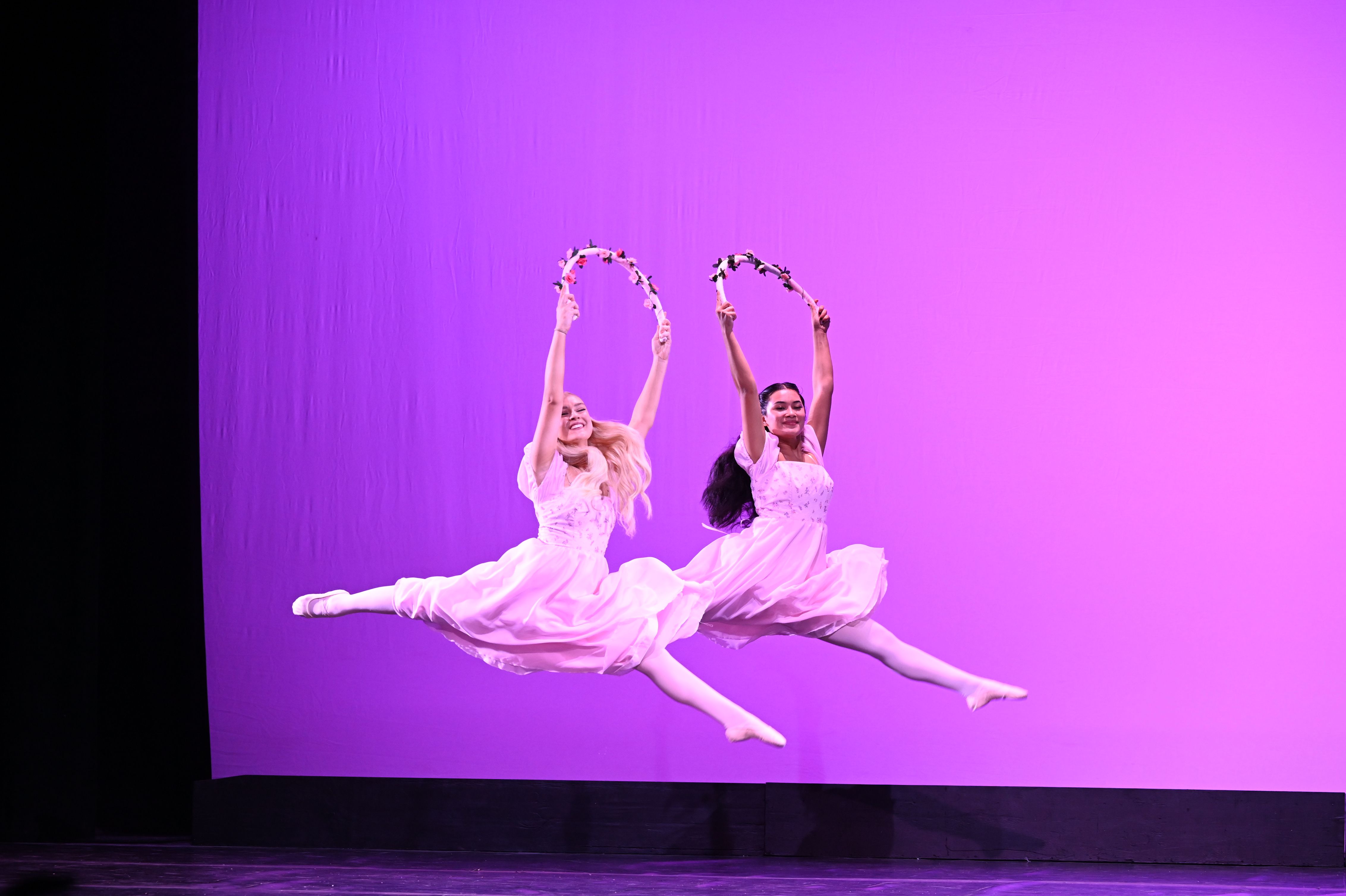 Two dancers leap together, each smiling and holding a rope adorned with flowers