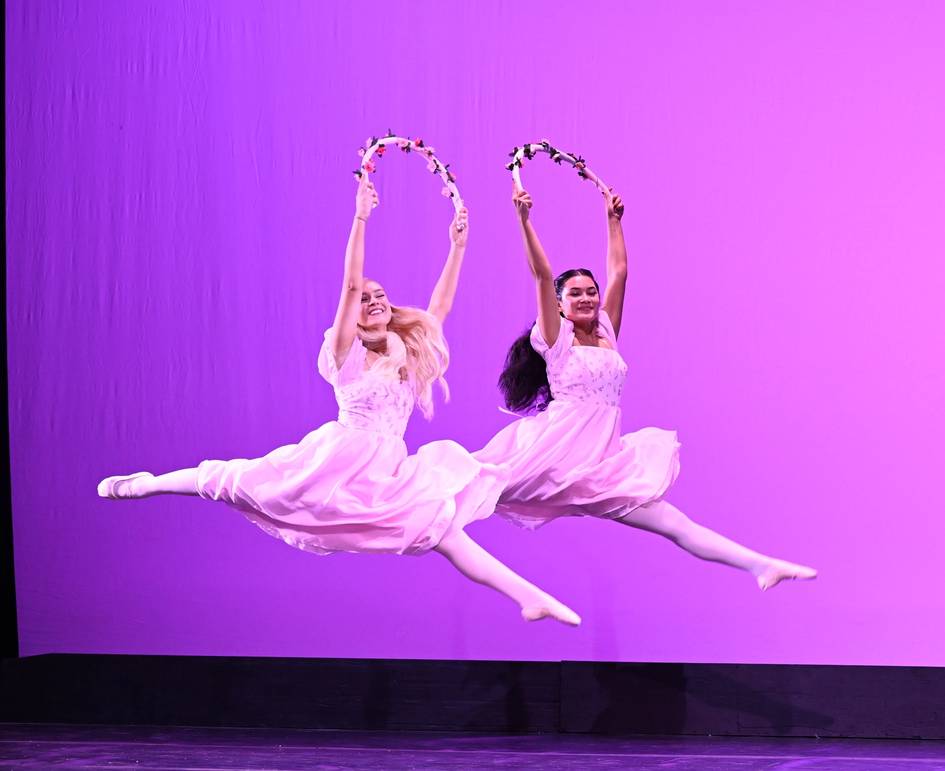 Two dancers leap together, each smiling and holding a rope adorned with flowers