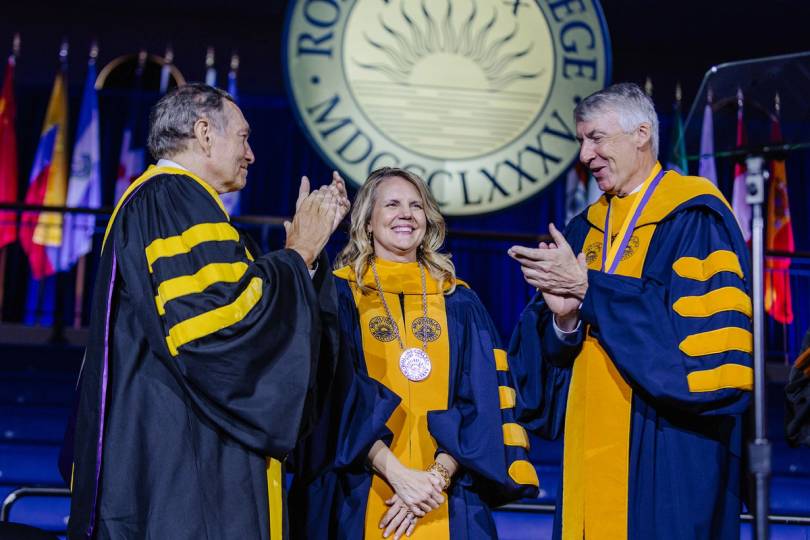 Rollins President Brooke Barnett stands on stage in academic regalia at the presidential inauguration with a college seal in the background.