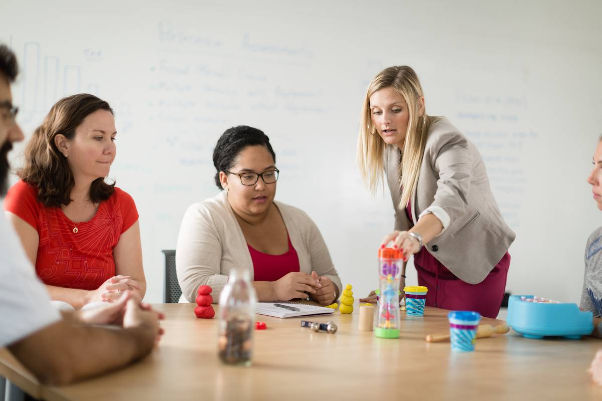 A small group of adults sits around a table as an instructor demonstrates a hands-on activity using colorful learning tools