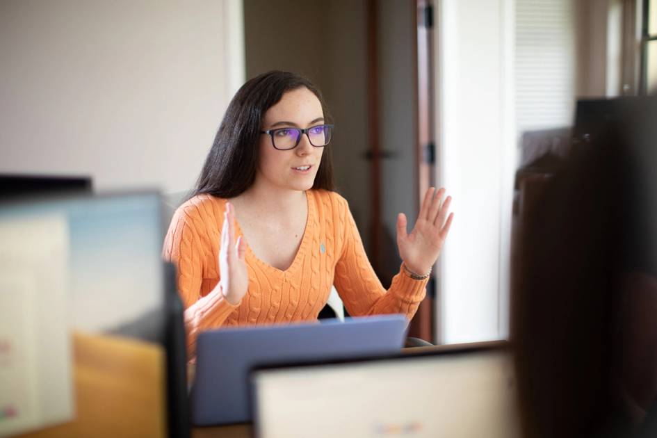 Person in orange sweater speaking animatedly during an office meeting