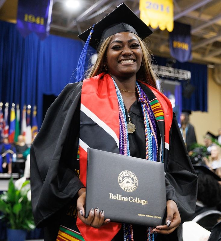 Student smiles at camera in cap and gown at commencement ceremony holding diploma