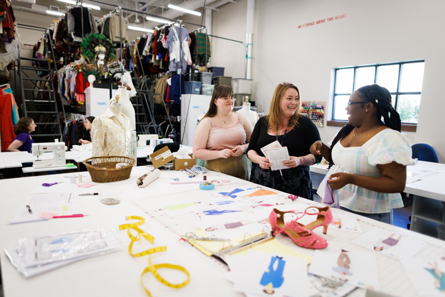 Two students on either side of a faculty member, talking happily at a drafting table in a costume studio with racks behind them