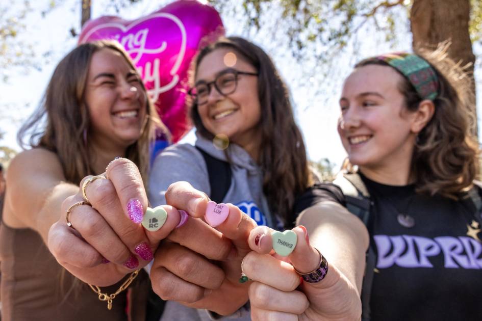 Three college girls holding candy hearts with I Heard Rollins, Fiat Lux, and Give Back on the hearts