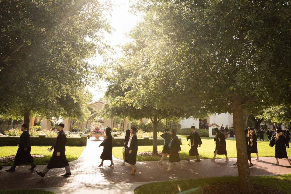 College students in caps and gowns walk across a sunlit college campus.