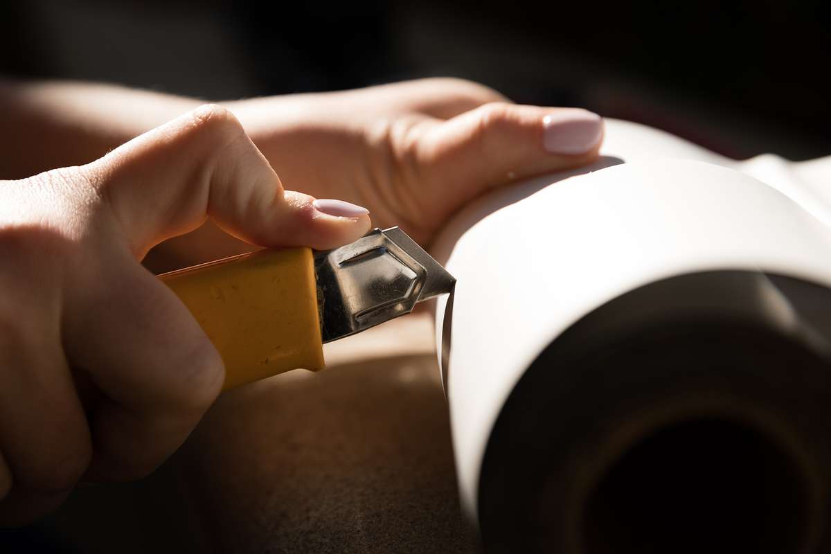 A close up picture of a student holding a razor knife cutting a roll of paper material.