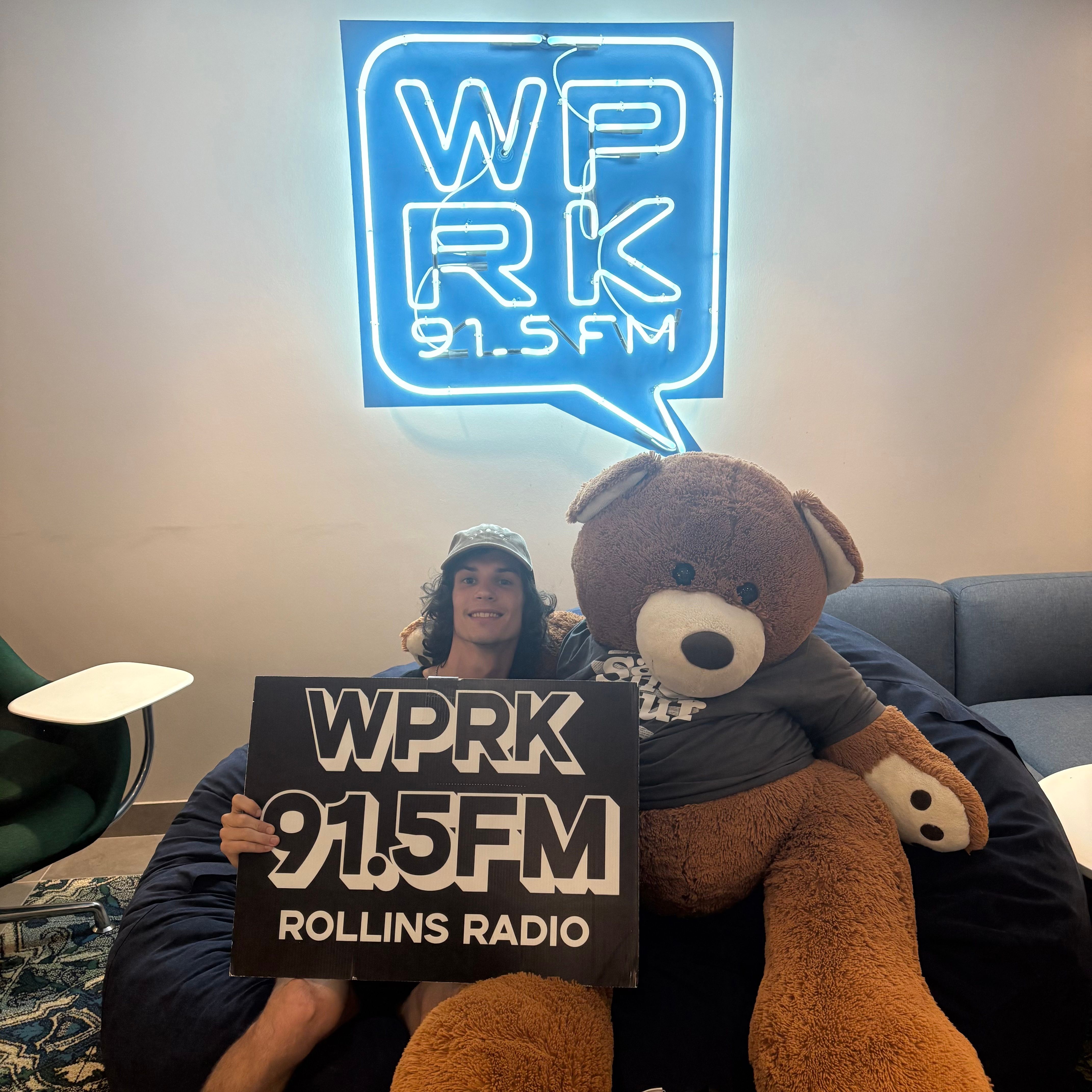 Student sits with a teddy bear holding a WPRK 91.5 FM Rollins Radio sign, in front of a WPRF 91.5 FM neon sign.