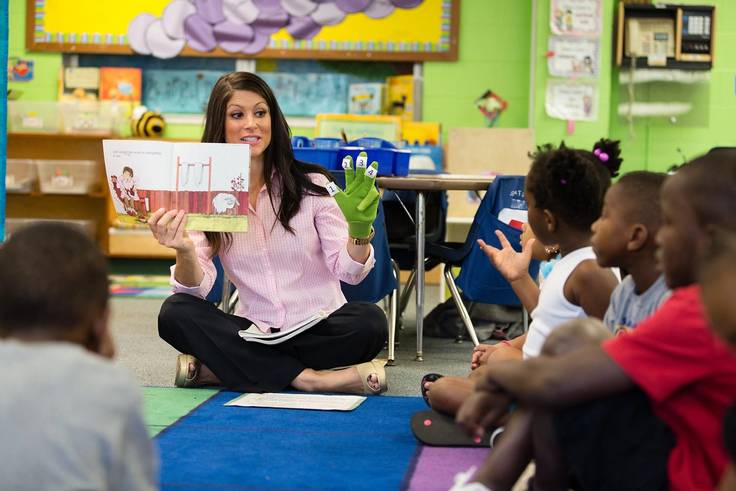 A Rollins education major is holding a book and a sock puppet, reading with an elementary class.