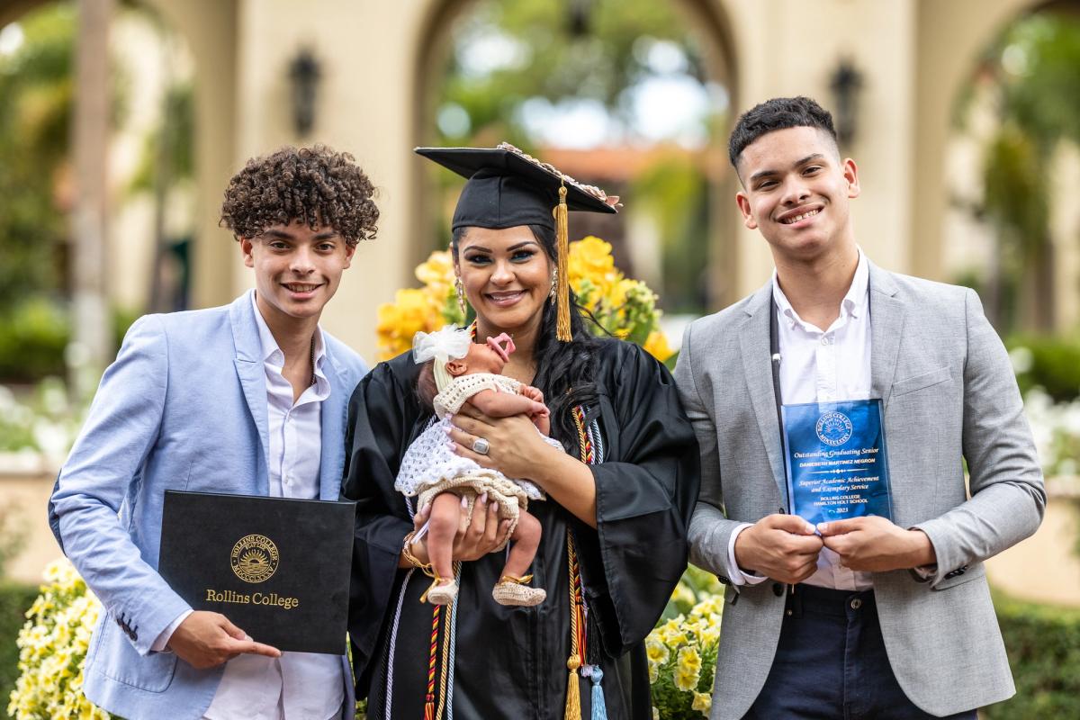 Female graduate holding baby girl, two young men holding diploma and award on either side.