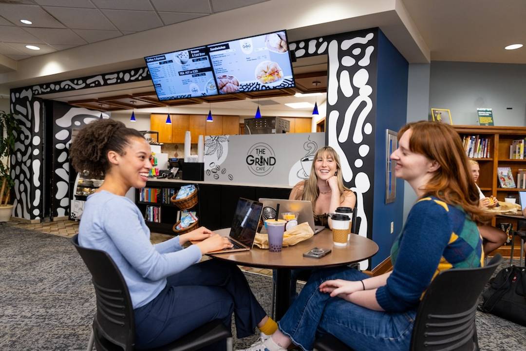 Three students sitting at a table, talking over coffee in the Olin Library in front of The Grind cafe.