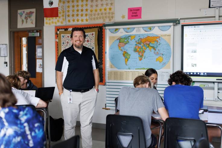 An alumni teacher stands in the center of a middle school classroom surrounded by his students.
