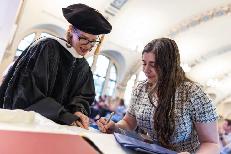 Professor in academic regalia assists Alessia Francesca Prenda ’26 signing document at PBK ceremony table.