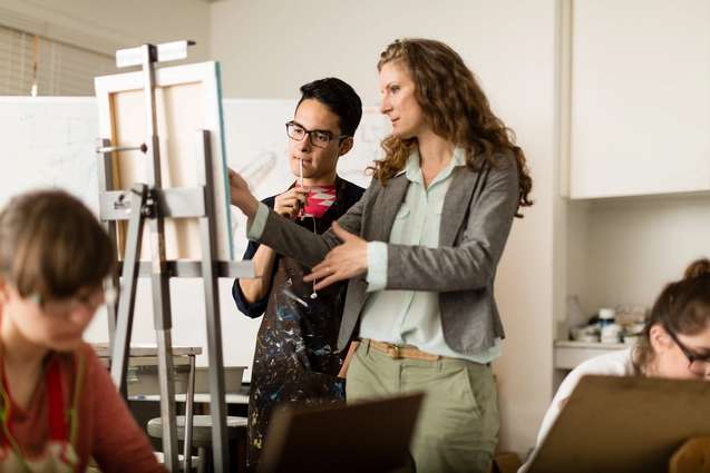 A studio art professor giving advice to her student as he paints his canvas resting on an easel.