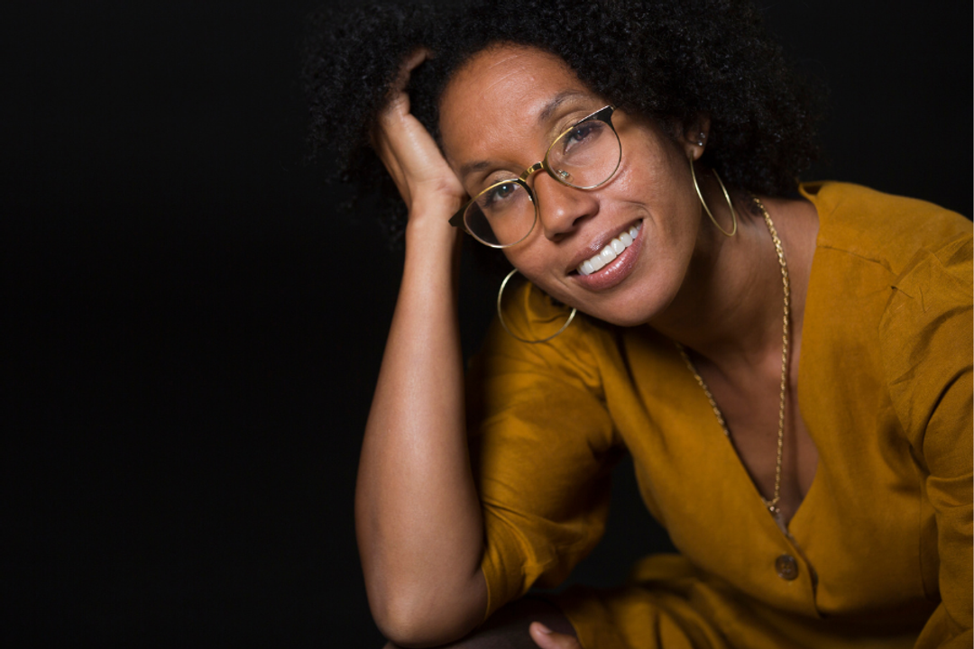 Smiling person wearing glasses and mustard clothing against black background
