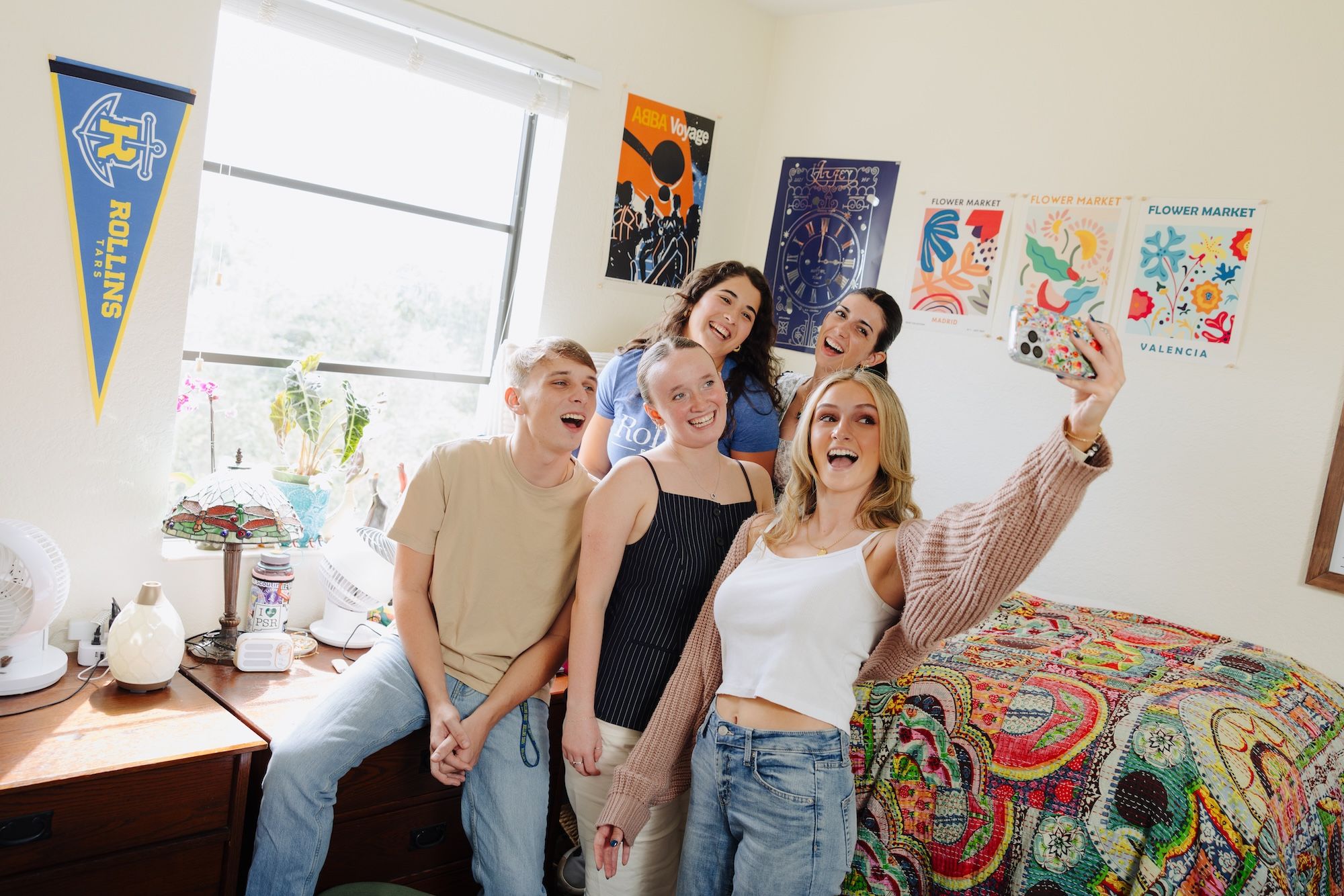 Five friends take a cheerful group selfie in a decorated bedroom.