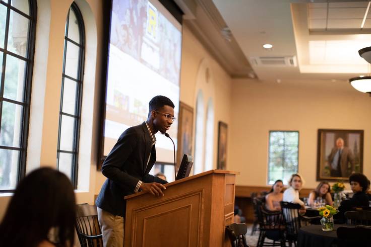 A college student delivers a talk at a podium to a small crowd of campus visitors.