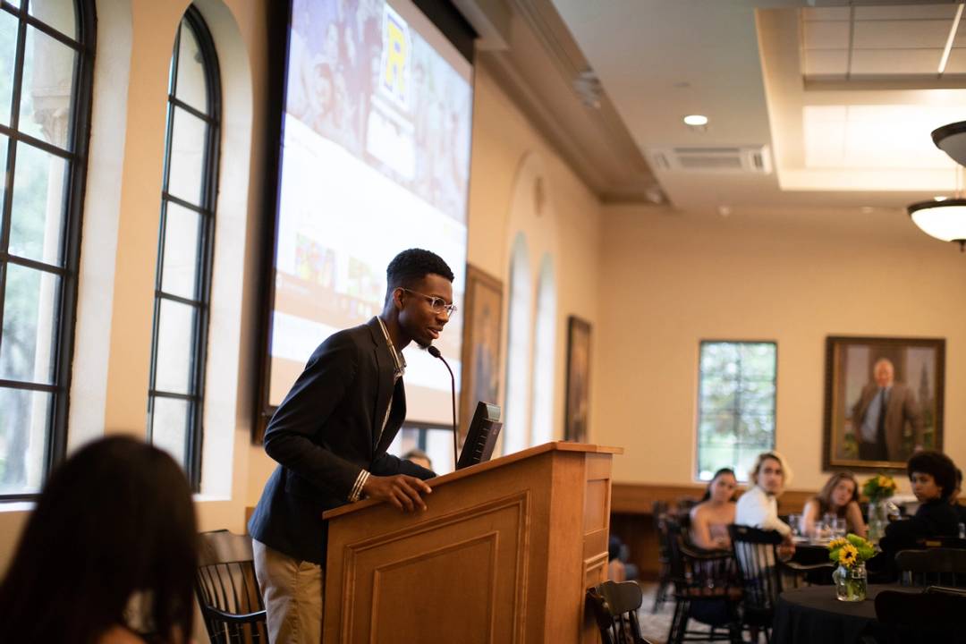 A college student delivers a speech at a podium to a group of high school students.