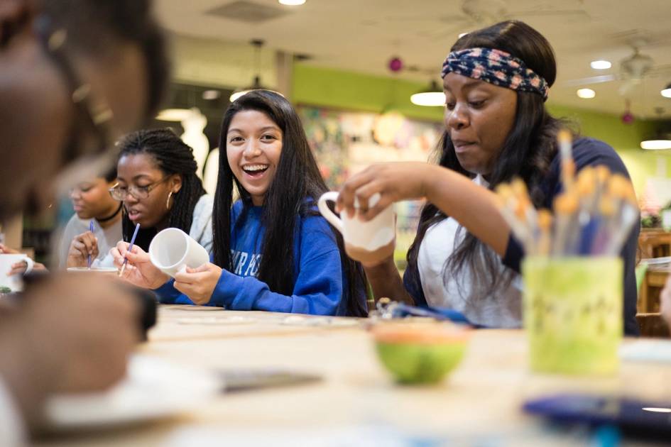 A college student smiles at the camera while participating in a mug decorating activity.