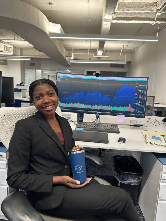 Smiling woman in a black suit sits at a desk holding a Unifi Asset Management tumbler, with a large monitor displaying a financial market chart behind her.
